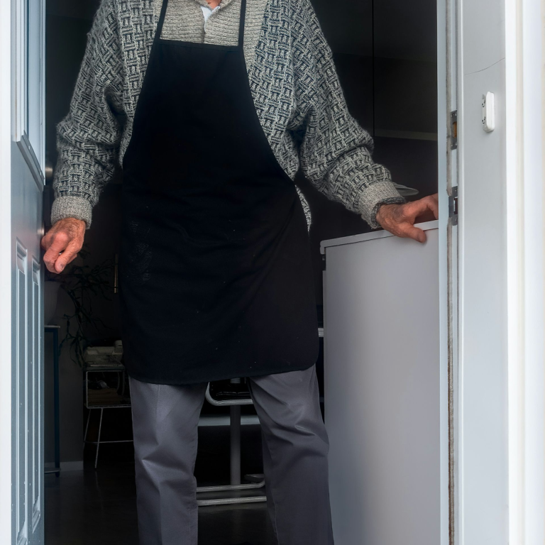 Haushalt Älterer Mann in Schürze steht an einer Tür, schaut freundlich nach draußen.